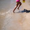 Bambina corre sulla spiaggia con un secchiello giallo.